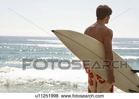 Stock Photo - Man holding surfboard by ocean back view. Fotosearch