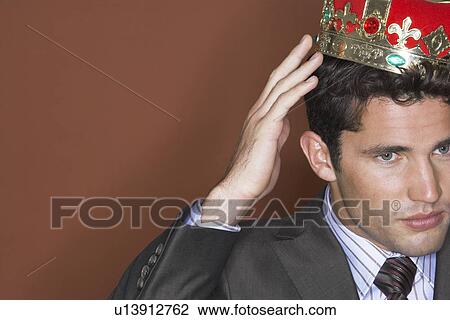 Stock Image - Man putting on crown against brown background close-up. Fotosearch