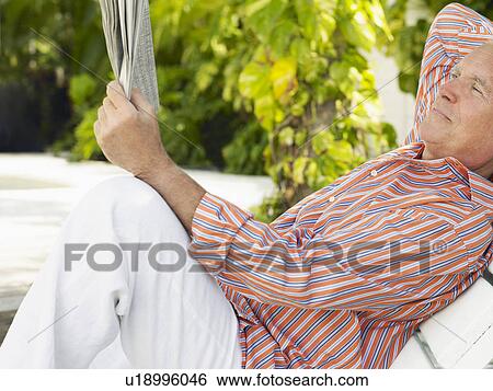 Stock Photograph - Man reclining on lounge chair reading newspaper side view. Fotosearch