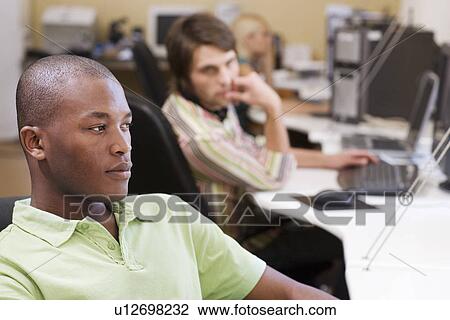 Stock Image - Man sitting at desk with man and woman in background using computers.. Fotosearch