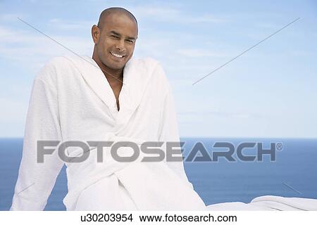 Picture - Man sitting in bathrobe smiling half length ocean behind. Fotosearch