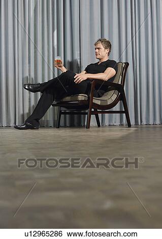 Man sitting in chair holding drink indoors View Large Photo Image Stock Photograph - Man sitting in chair holding drink indoors. Fotosearch