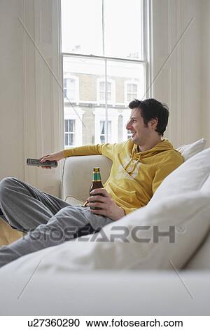 Stock Image - Man sitting on sofa watching television and holding beer bottle. Fotosearch
