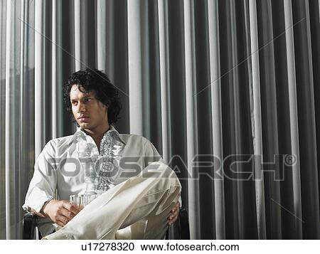 Stock Image - Man sitting with alcoholic drink in front of curtains. Fotosearch