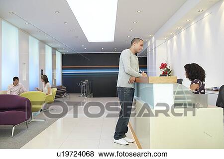 Stock Photo - Man standing at reception desk Talking to Receptionist side view. Fotosearch