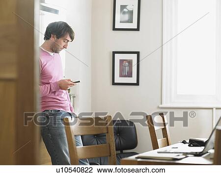 Stock Image - Man standing in kitchen text messaging. Fotosearch