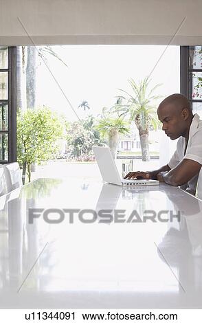 Stock Image - Man using laptop on dining table side view. Fotosearch