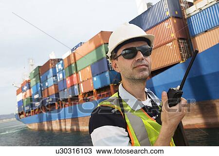 Stock Image - Man wearing hard hat using walkie-talkie at container terminal. Fotosearch