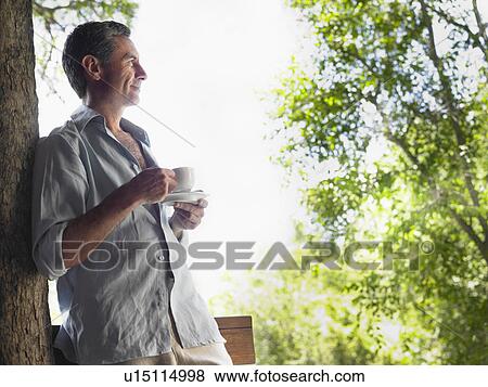 Stock Photo - Mature man drinking tea leaning on tree looking at view profile. Fotosearch