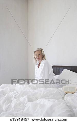 Stock Photo - Mature woman in bathrobe sitting up in bed smiling next to open book side view. Fotosearch