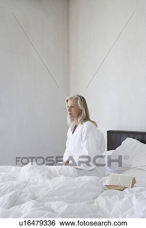 Stock Photograph - Mature woman in bathrobe sitting up in bed next to open book side view. Fotosearch
