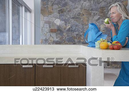 Stock Image - Mature woman leaning on countertop eating apple side view. Fotosearch
