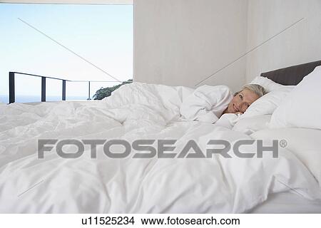 Picture - Mature woman lying in bed smiling. Fotosearch