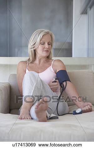 Stock Photo - Mature woman sitting on sofa barefoot checking blood pressure with sphygmometer. Fotosearch