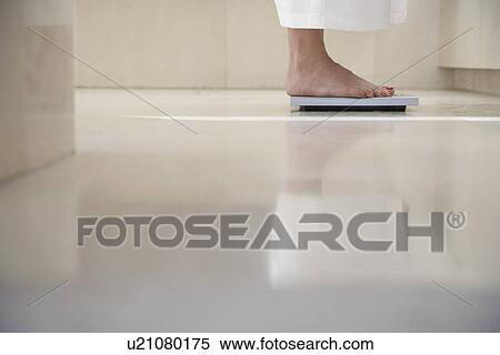Stock Photography - Mature woman standing on scale in bathroom weighing herself low section. Fotosearch