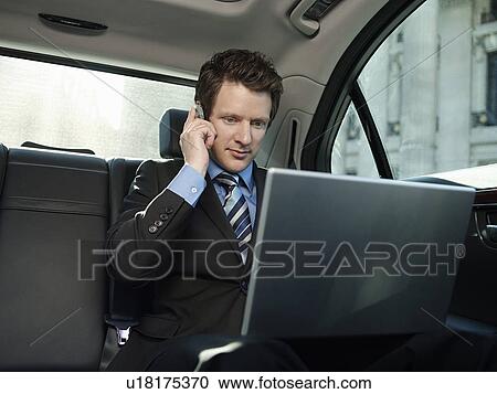 Stock Image - Mid adult businessman sitting at back seat of car using laptop and mobile phone. Fotosearch