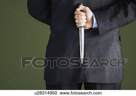 Stock Image - Mid-adult businessman standing holding knife behind back mid section back view. Fotosearch