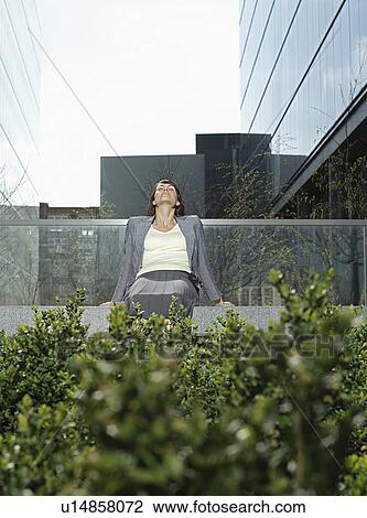 Stock Image - Mid adult businesswoman relaxing on wall outside office buildings. Fotosearch