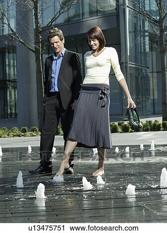 Stock Image - Mid-adult couple walking between water jets of fountain. Fotosearch