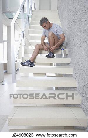 Stock Image - Mid-adult man sitting on stairs tying shoes. Fotosearch