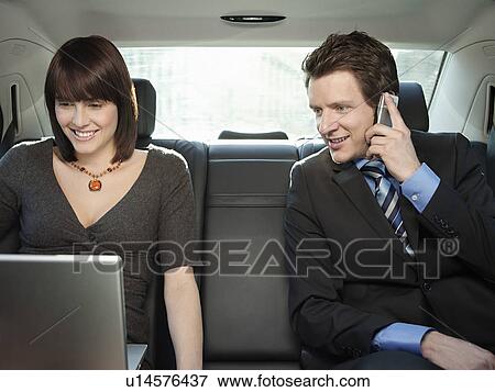 Stock Photo - Mid adult woman and businessman sitting at back seat of car using mobile phone and laptop. Fotosearch