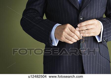Picture - Middle-aged businessman in full suit holding pair of glasses mid section. Fotosearch