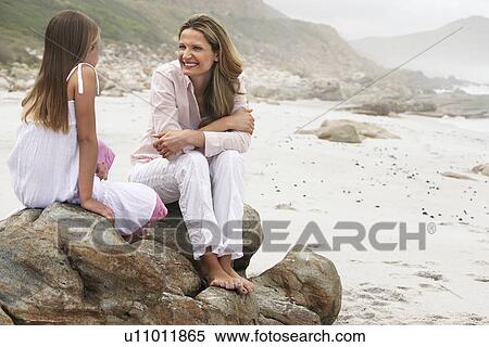Stock Photography - Mother and daughter (10-12) sitting on boulder on beach. Fotosearch