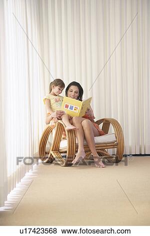 Stock Photo - Mother and daughter (5-6) sitting in armchair reading book. Fotosearch