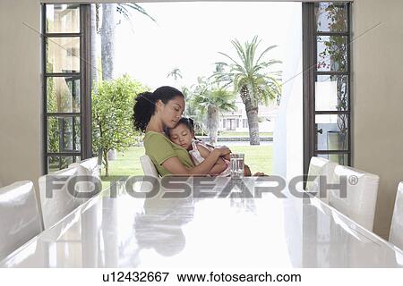 Stock Photo - Mother and daughter (5-6 years) embracing sitting at dining table. Fotosearch