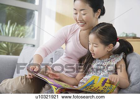 Stock Image - Mother sitting with daughter on sofa reading a book. Fotosearch