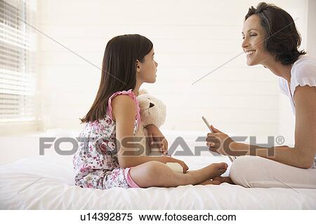 Stock Photography - Mother using lap top daughter cuddling teddy bear sitting face to face on bed. Fotosearch