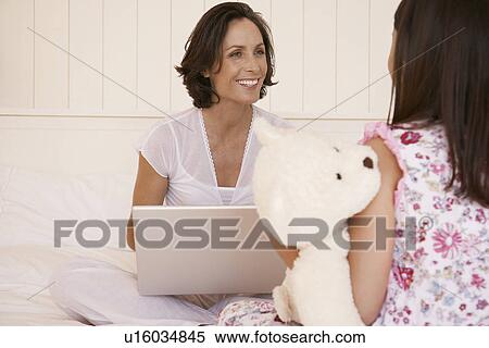 Mother using lap top daughter cuddling teddy sitting on bed View Large Photo Image Stock Photography - Mother using lap top daughter cuddling teddy sitting on bed. Fotosearch