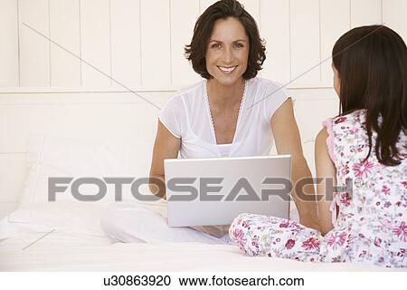Stock Image - Mother using lap top sitting with daughter on bed. Fotosearch