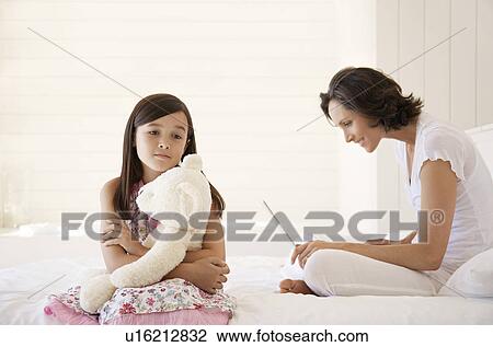 Stock Image - Mother using laptop daughter cuddling teddy sitting on bed. Fotosearch