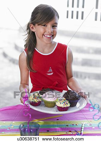 Stock Image - Portrait of girl (7-9) holding tray with cupcakes smiling. Fotosearch