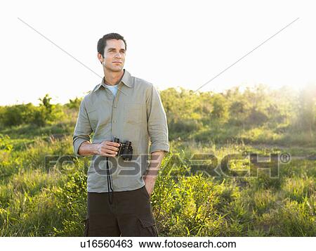 Stock Image - Portrait of mid adult man in fields holding binoculars. Fotosearch