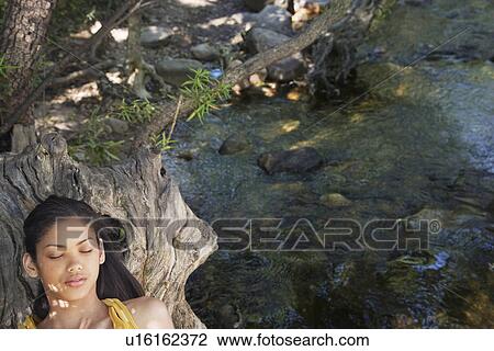 Stock Image - Portrait of teenage girl (16-17 years) relaxing on tree trunk by stream. Fotosearch
