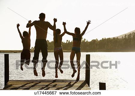 Stock Photo - Rear view of father and children jumping off a pier holding hands. Fotosearch