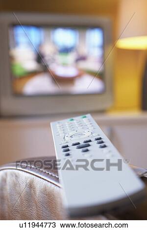 Stock Image - Remote control lying on armchair with television turned on in background close-up. Fotosearch