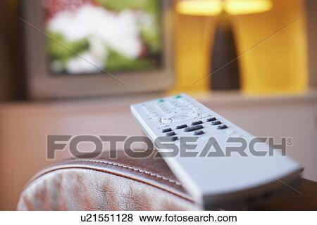 Stock Photo - Remote control lying on armchair with television turned on in background close-up. Fotosearch