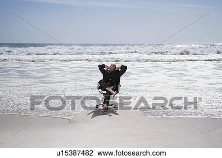Stock Image - Senior business man sitting on office chair on beach elevated view. Fotosearch