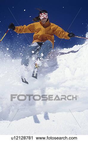 Stock Image - Skier skiing through snow jumping from snow bank. Fotosearch