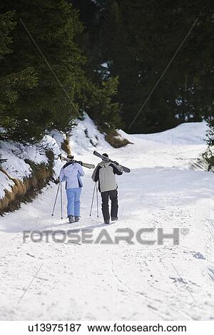 Stock Photo - Skiing couple walking down ski slope carrying skis on shoulders back view elevated view. Fotosearch