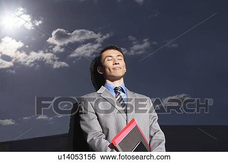 Stock Photograph - Smiling Businessman leaning against reflective wall outside eyes closed holding notebook. Fotosearch