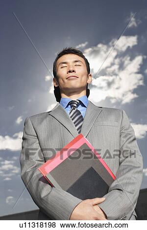 Stock Photo - Smiling Businessman standing with eyes closed holding notebook low angle view. Fotosearch