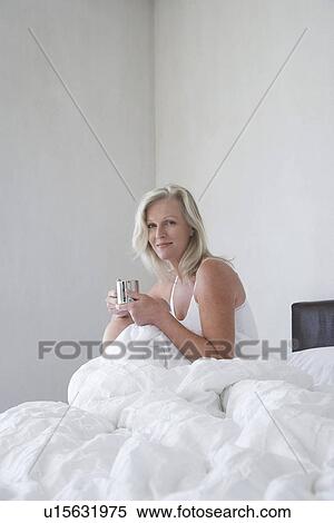 Stock Photography - Smiling Mature woman in pyjamas holding cup sitting up in bed side view. Fotosearch
