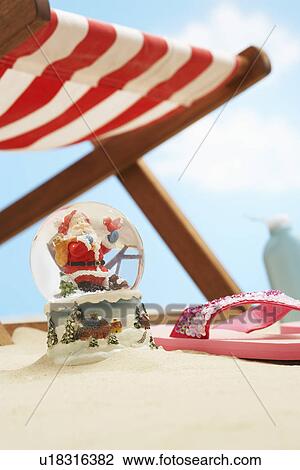 Stock Image - Souvenir santa snow globe under deckchair on beach close up. Fotosearch
