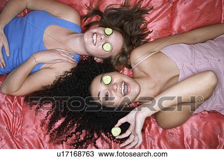 Stock Image - Teenage Girls lying on backs on pink sheets with cucumbers in eyes overhead view. Fotosearch