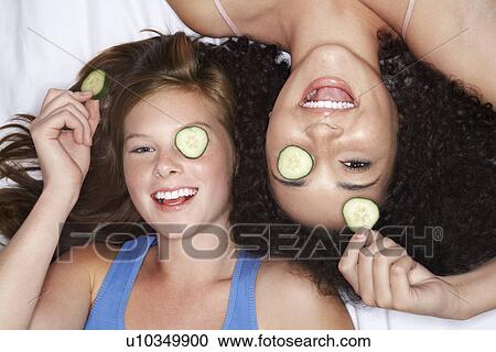 Stock Image - Teenage Girls lying on backs with cucumbers in eyes overhead view. Fotosearch