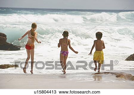 Stock Image - Three children (5-6 7-9 10-12) running on beach back view. Fotosearch